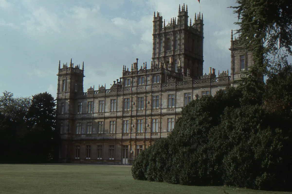 The towered stone façade of Highclere Castle beneath a grey sky, half screened by dark evergreens